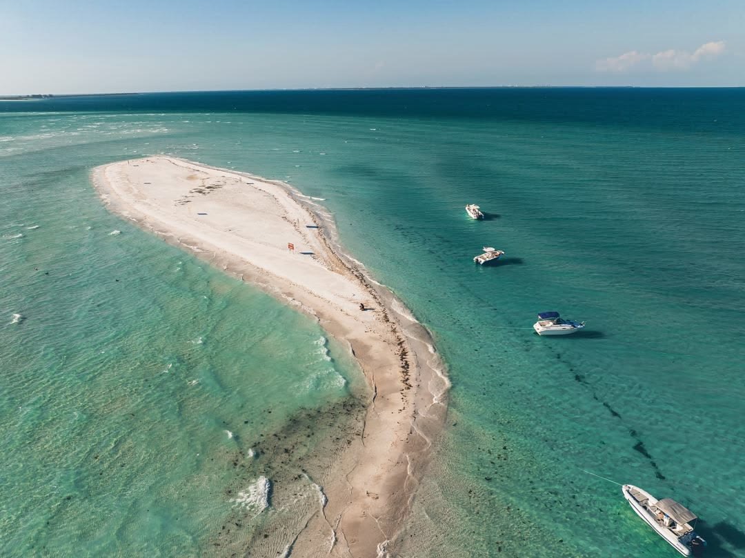 Aerial view of a curved white sandbar surrounded by clear turquoise Gulf water with small boats anchored nearby